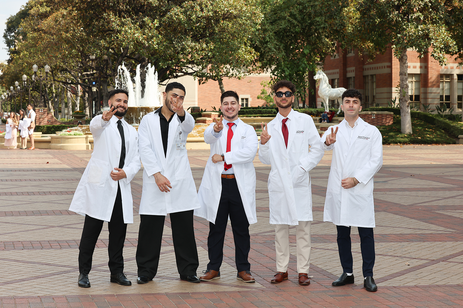 Five dental students with white coats on flashing a Fight On sign in front of Traveler on USC Campus